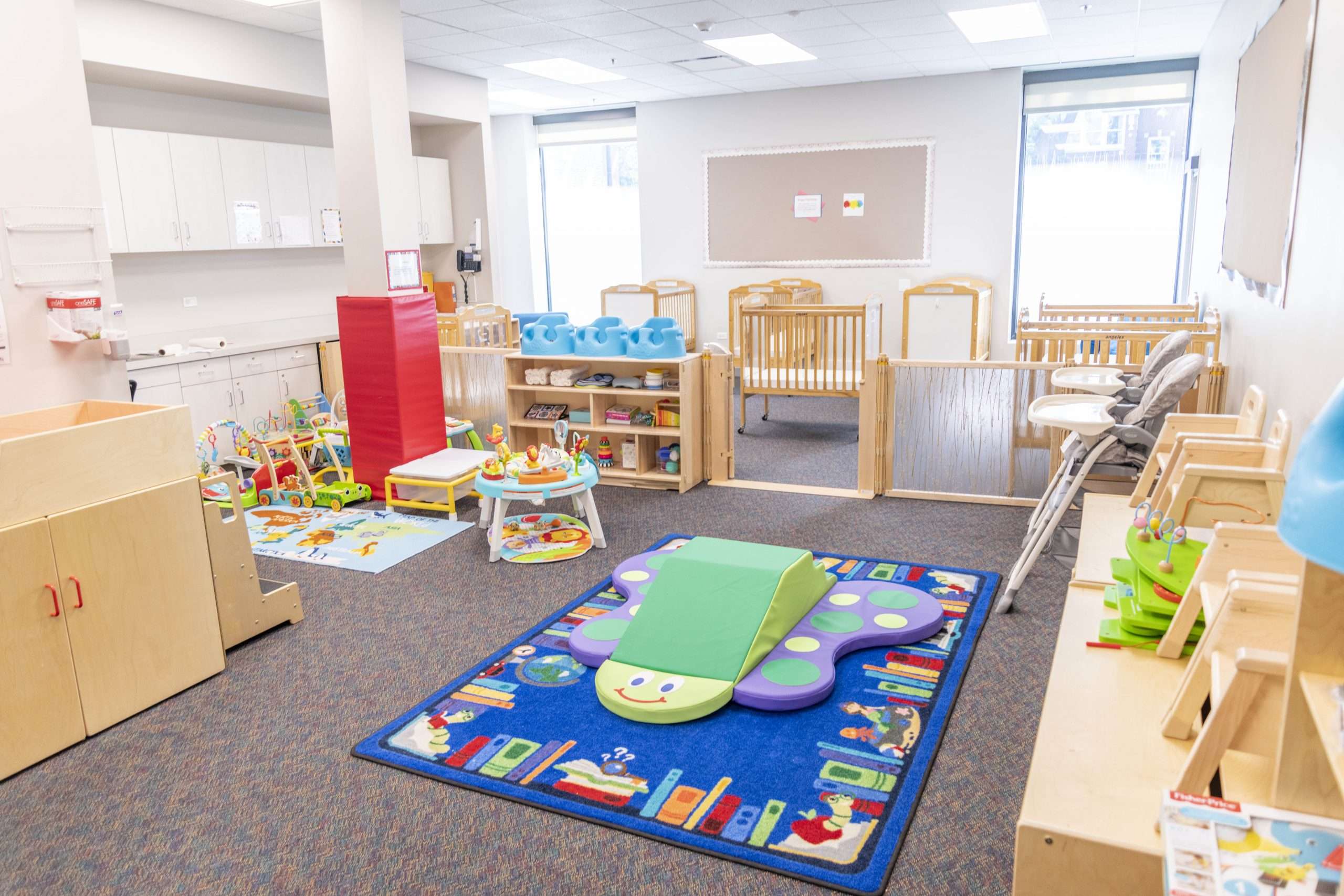 Brightly lit carpeted infant classroom at JCYS Lincoln Square. Wooden cabinets located on the each side of the room, a blue rug and pastel climber in the middle of the room. Cribs and sleeping area are in the back of room.