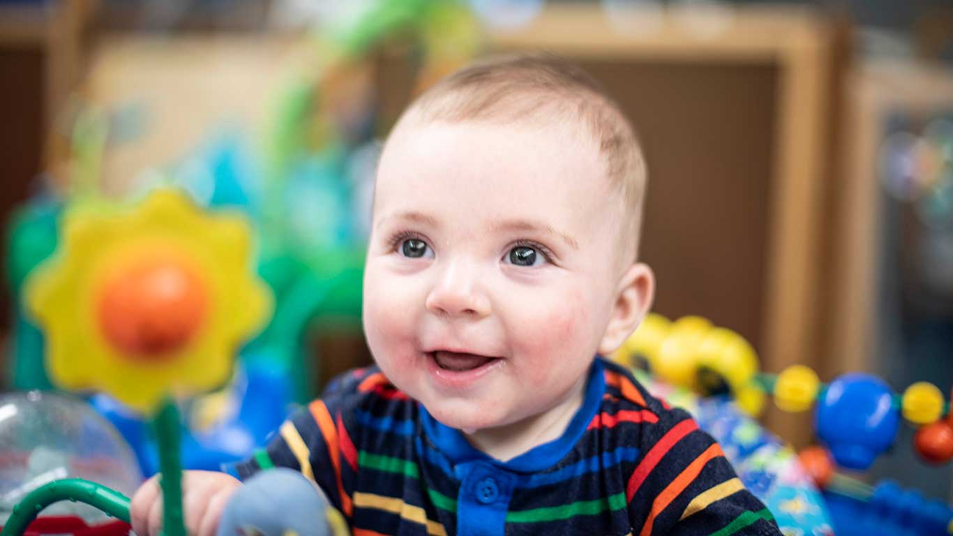 Infant in colorful onesie smiling and looking at a toy flower with yellow petals, an orange center, and green stem. Links to JCYS Highland Park infant care page.