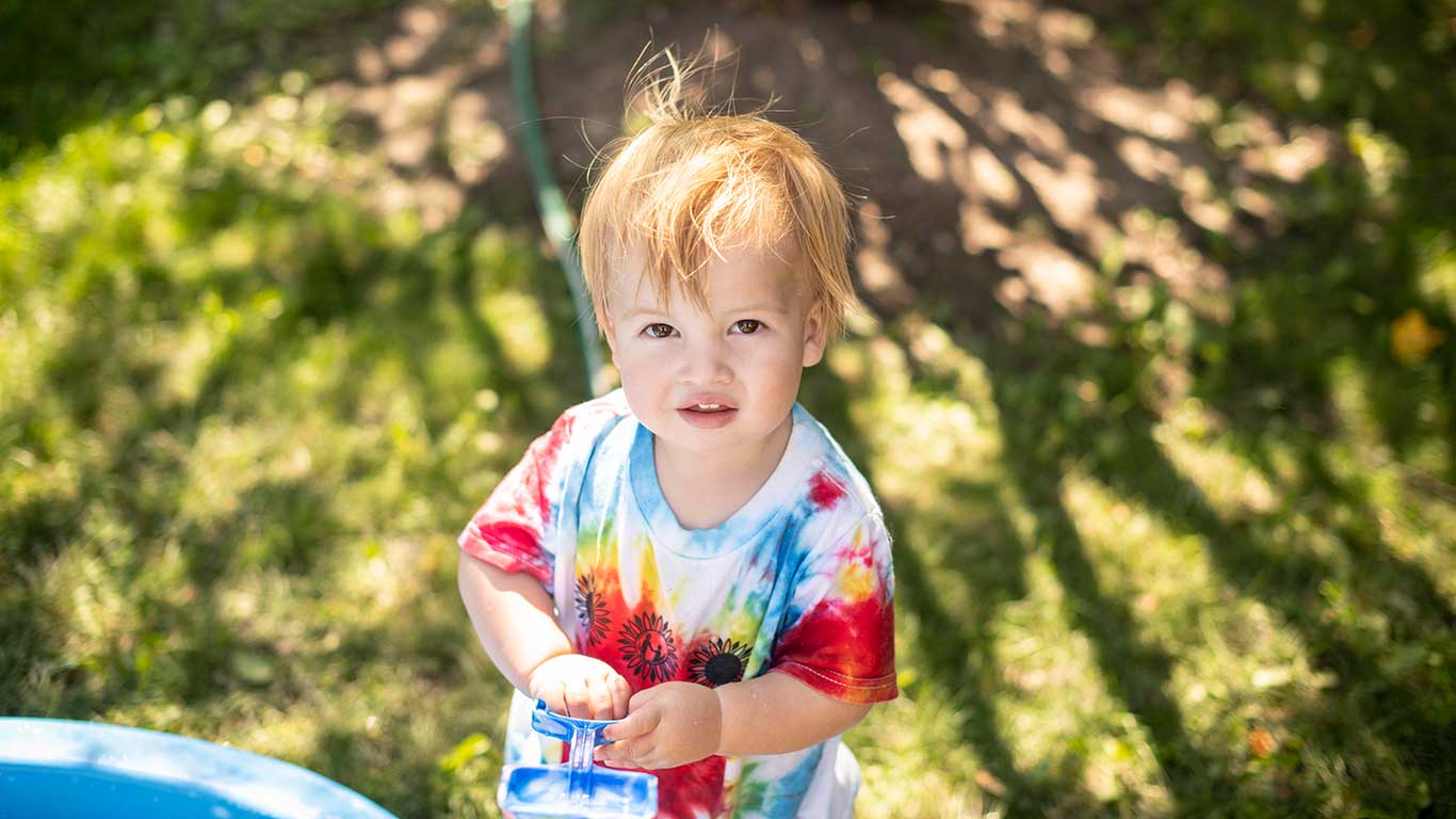 Toddler standing in the shade of a tree. Toddler is wearing tie-dyed Sunflower Camp t-shirt looking up and smiling at the camera.