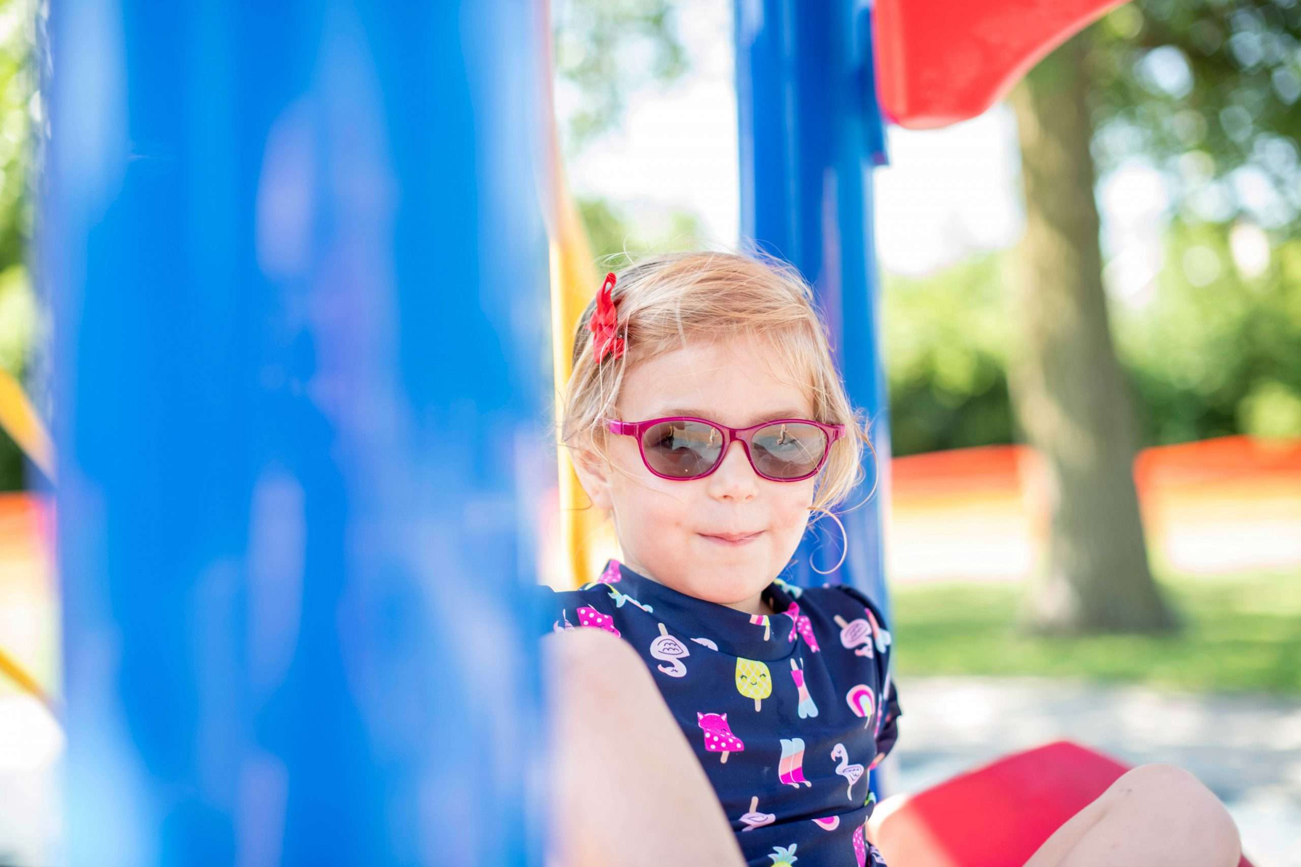Child sitting on playground equipment. They are wearing pink sunglasses and looking directly at the camera with a smirk on their face.