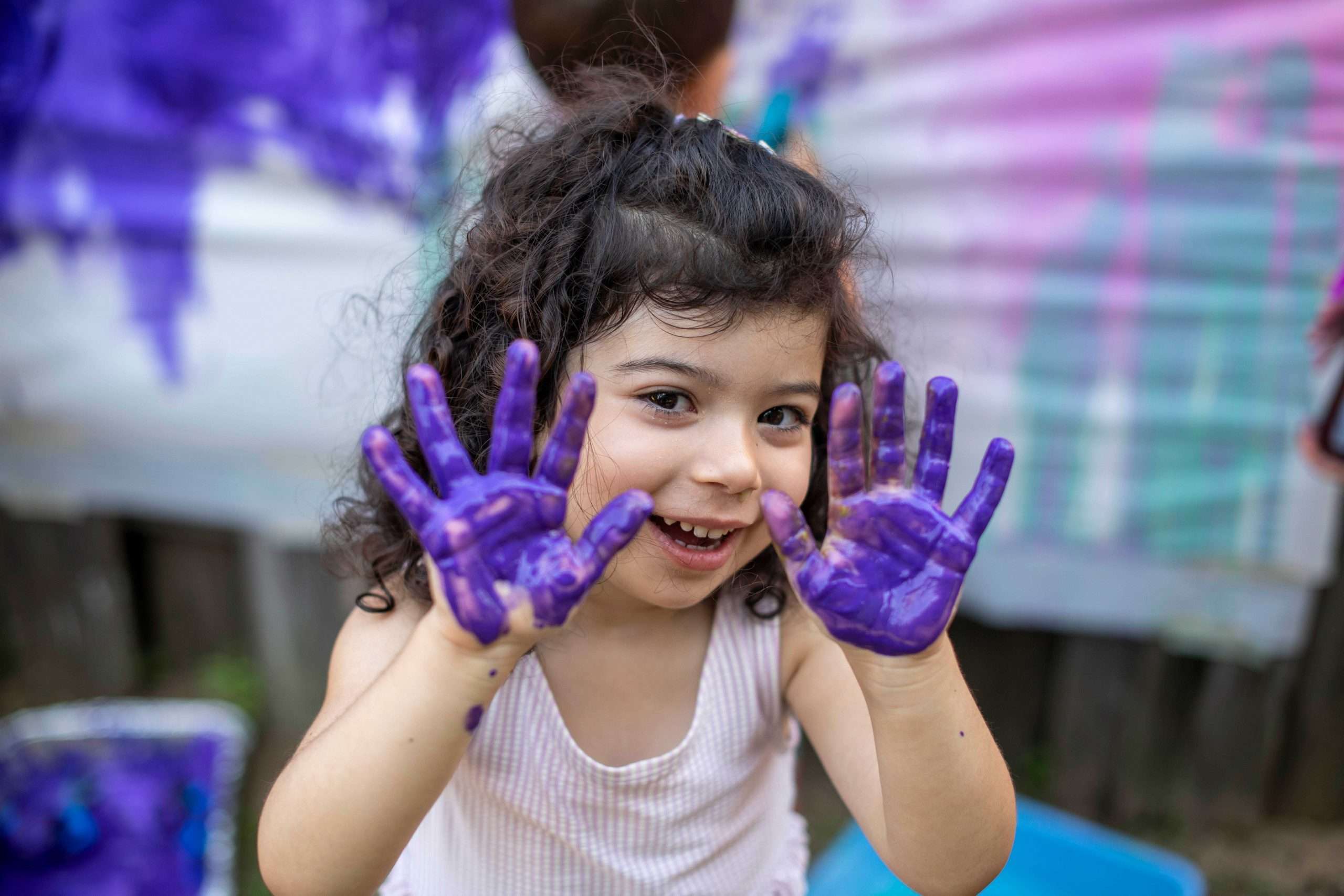 Child smiling in front of finger painted canvas. Child is showing both of their hands which are covered in purple paint.