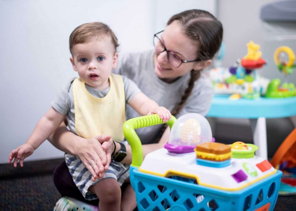 Teacher is sitting on floor and smiling at the infant they are holding. The infant is looking at the camera and holding on to a play shopping cart