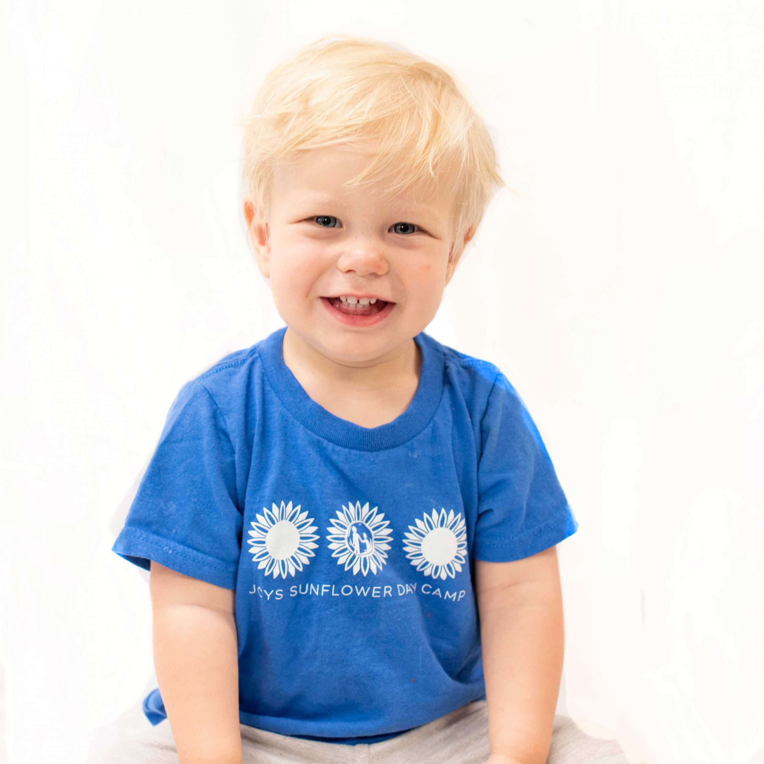 Boy looking at camera and smiling, wearing a blue JCYS Sunflower Day Camp shirt.